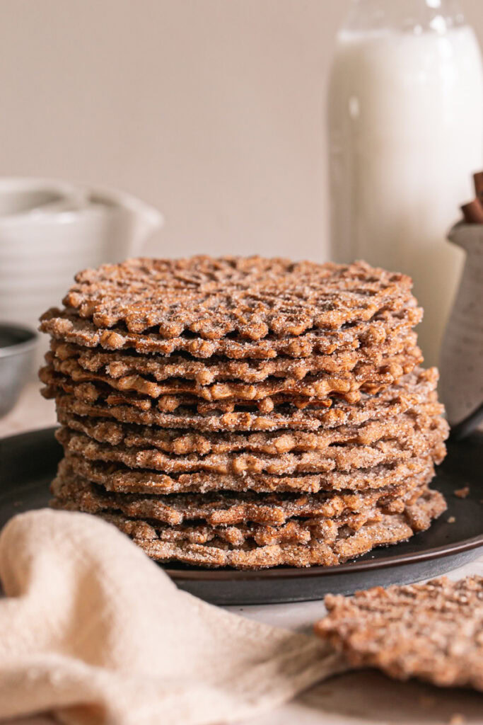 a close up of a stack of cinnamon sugar pizzelles on a round platter with handles, a jar of milk, a jug of cinnamon sticks and a linen napkin