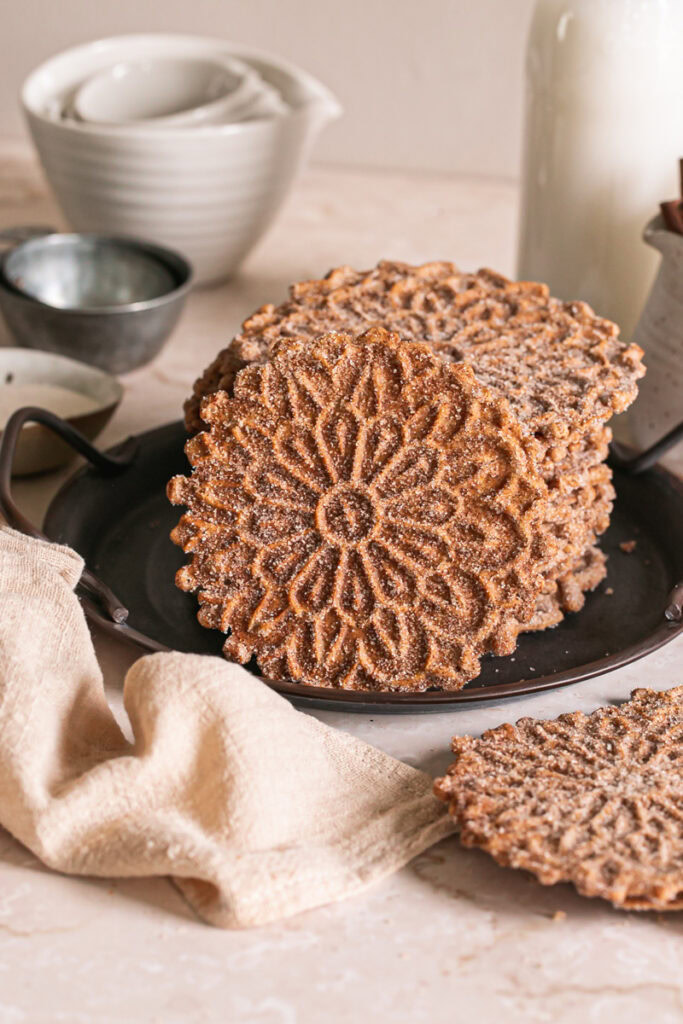 a stack of cinnamon sugar pizzelles, one leaning against the side of the stack to reveal the pizzelle pattern on the top