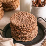 a stack of cinnamon sugar pizzelles on a round platter with handles, a jar of milk, a jug of cinnamon sticks and a linen napkin