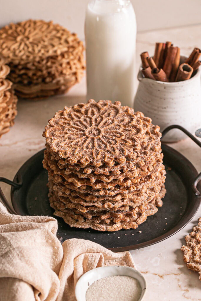 a stack of cinnamon sugar pizzelles on a round platter with handles, a jar of milk, a jug of cinnamon sticks and a linen napkin