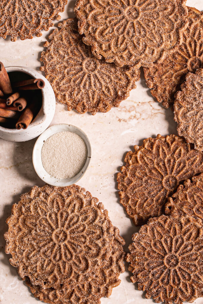 Cinnamon Sugar Pizzelles laid out on a marble backdrop with a ceramic cup filled with cinnamon sticks and a small pinch bowl filled with sugar