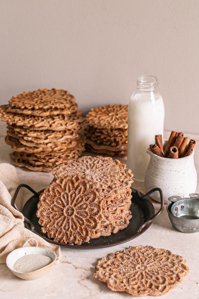 a stack of cinnamon sugar pizzelles, one leaning against the side of the stack to reveal the pizzelle pattern on the top and a stack of unsugared pizzelles in the background