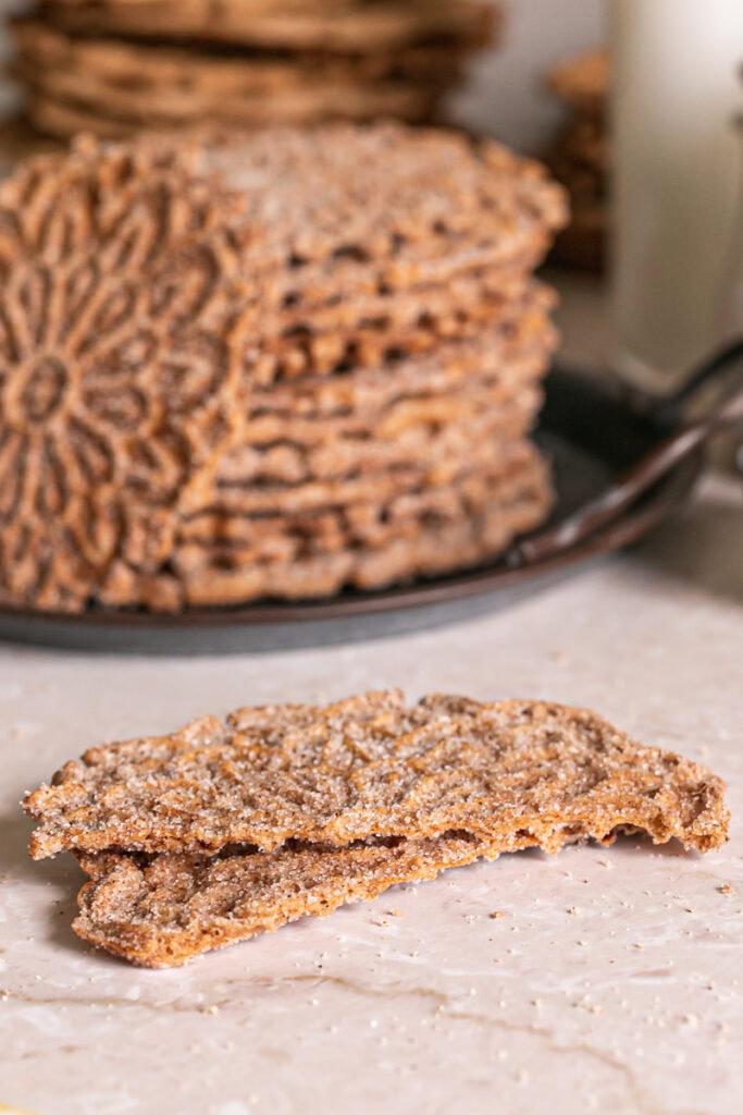 a cinnamon sugar pizzelle broken in half laying in front of a stack of pizzelles