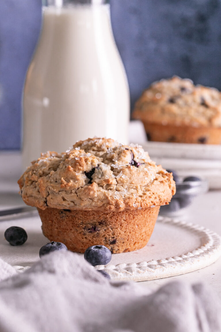 one blueberry muffin on a plate with a jar of almond milk in the background and another muffin on a stack of plates with fresh blueberries around it