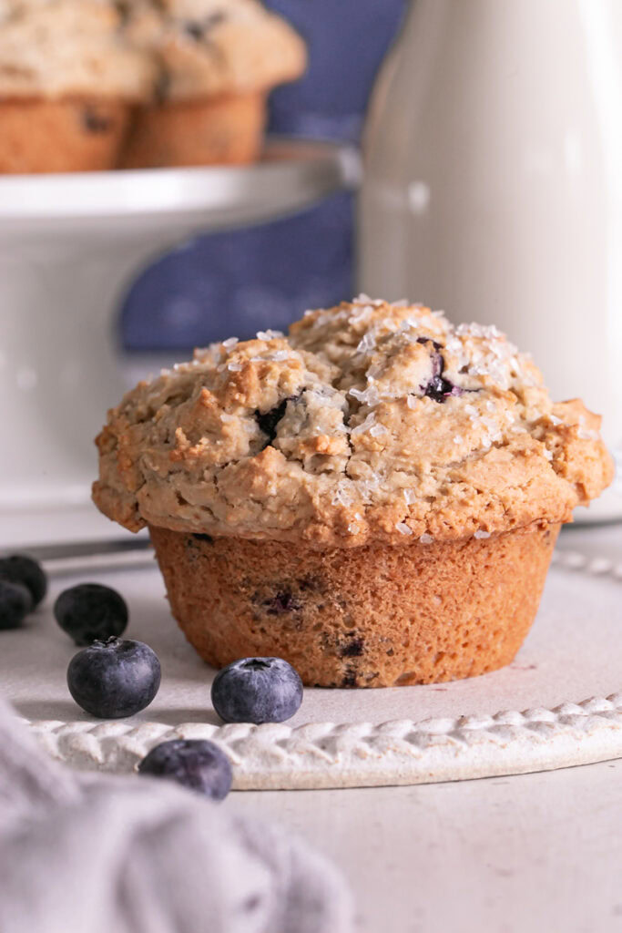 close up of a blueberry muffin on a plate with a jar of almond milk in the background and another muffin on a stack of plates with fresh blueberries around it