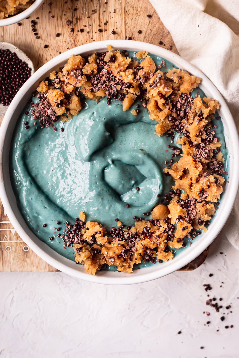 a blue spirulina smoothie bowl topped with almond flour cookie dough, chocolate covered chia seeds on top of a wooden cutting board with a bowl of cookie dough and cacao nibs beside it
