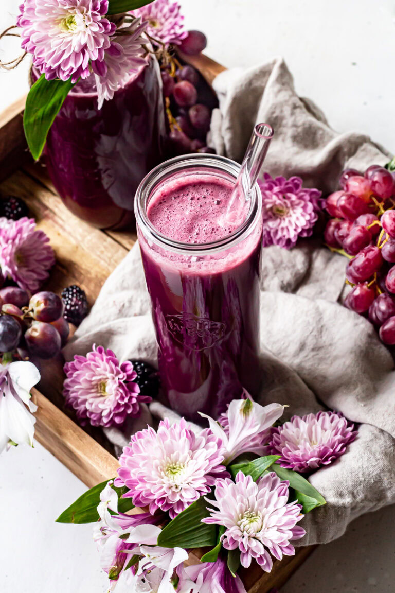 a glass of purple passion blender juice with purple flowers and grapes around it.