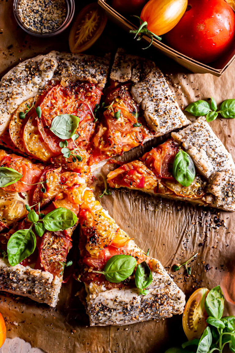 close up of a sliced tomato galette with a basket of tomatoes beside it and fresh basil leaves