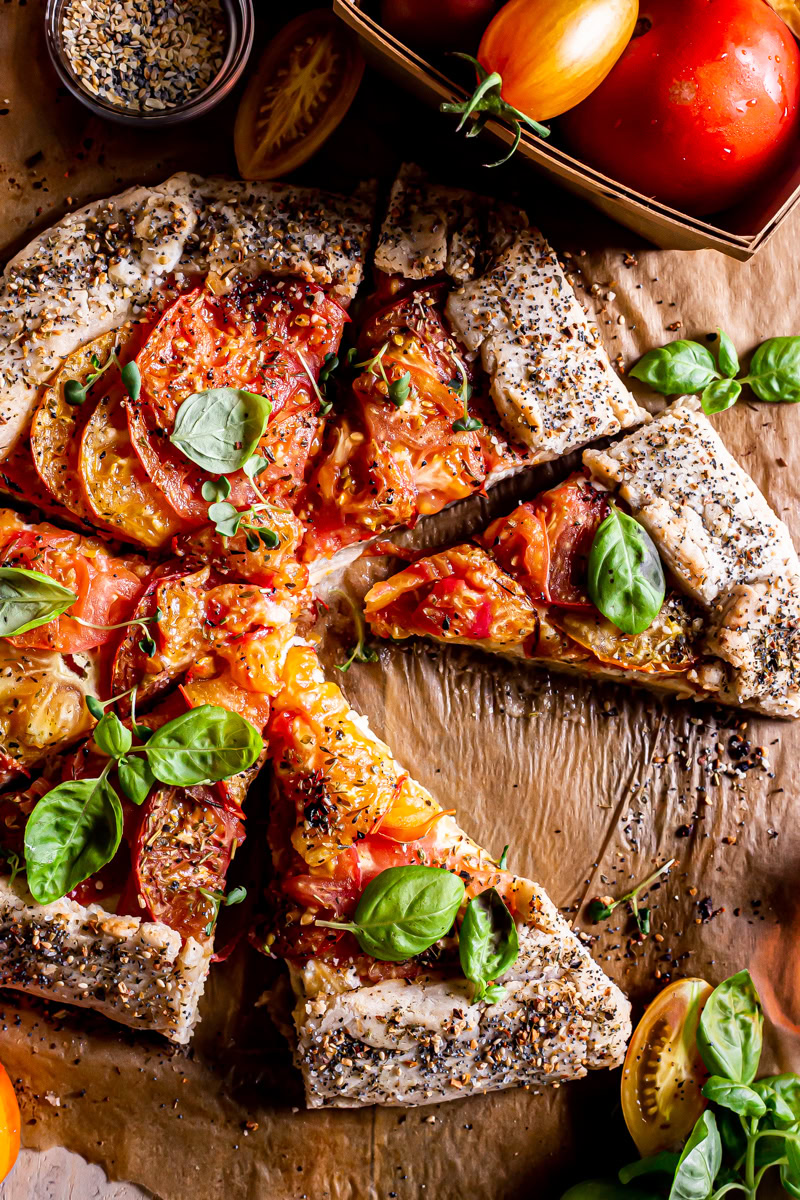 close up of a sliced tomato galette with a basket of tomatoes beside it and fresh basil leaves