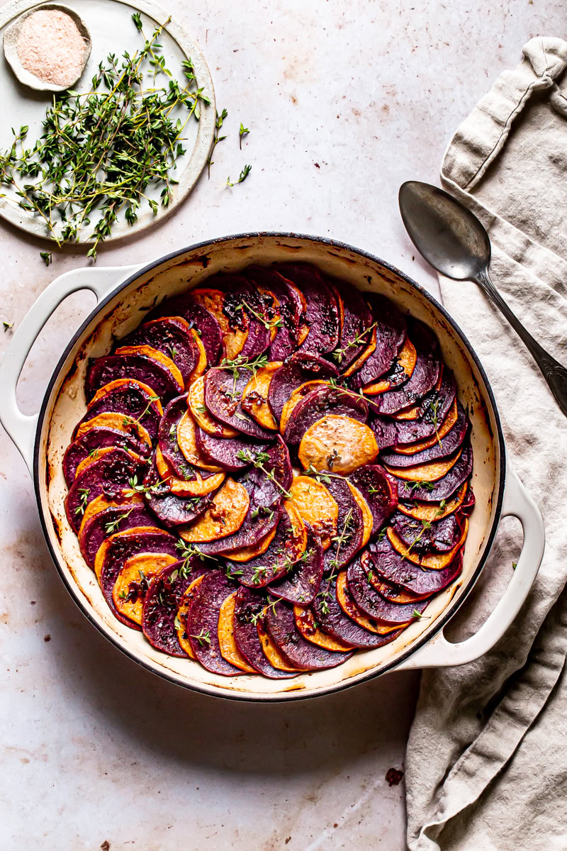 a round skillet with roasted purple and orange sweet potatoes in a colored pattern with fresh thyme and linen napkin and spoon on the side