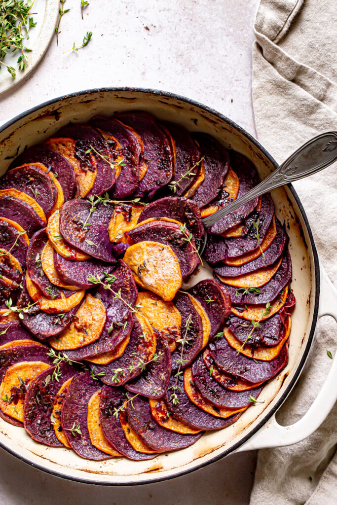 a round skillet with roasted purple and orange sweet potatoes in a colored pattern with fresh thyme and linen napkin and spoon scooping out some potatoes