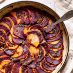 a round skillet with roasted purple and orange sweet potatoes in a colored pattern with fresh thyme and linen napkin and spoon scooping out some potatoes