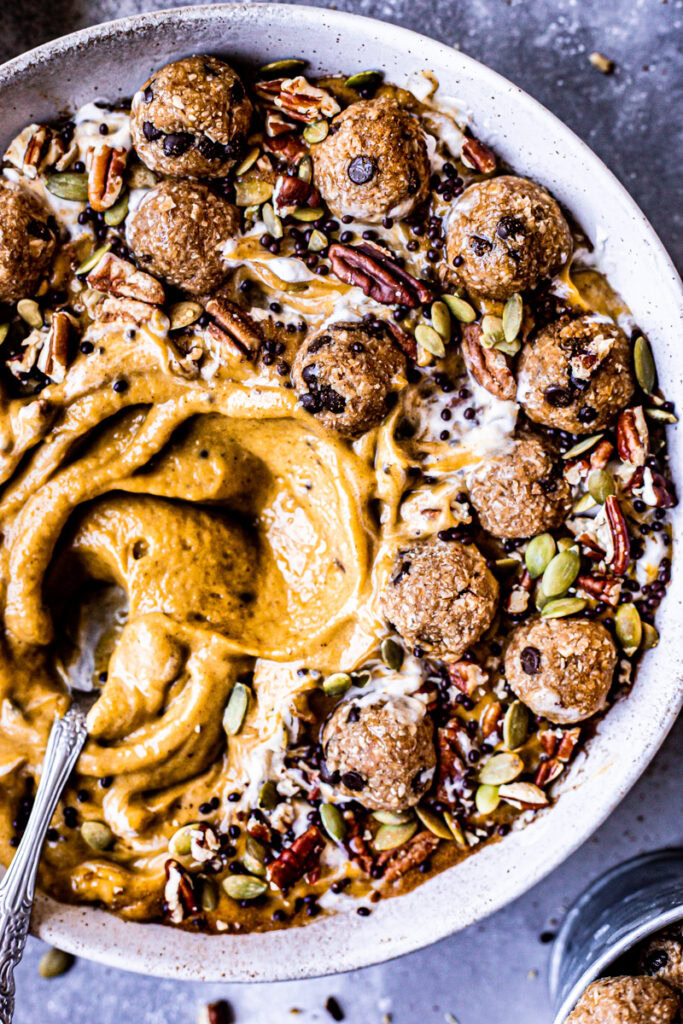 a close up of a pumpkin pie smoothie in a bowl topped with cookie dough balls, pumpkin seeds, pecans, chocolate covered chia seeds drizzled with maple syrup