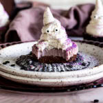 vegan cheese cake with a chocolate crust and purple vegan cashew cheesecake layer topped with a buttercream frosting ghost with candy eyes and purple sprinkles.