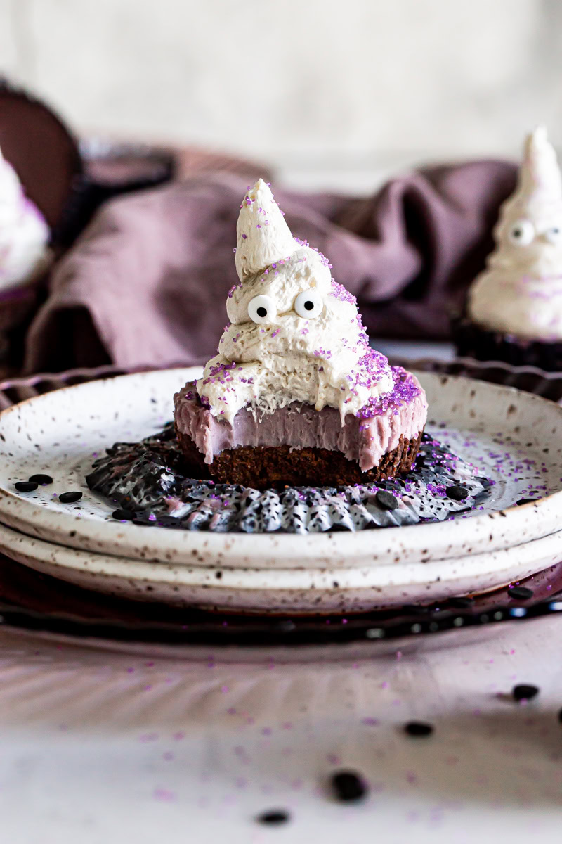 vegan cheese cake with a chocolate crust and purple vegan cashew cheesecake layer topped with a buttercream frosting ghost with candy eyes and purple sprinkles.