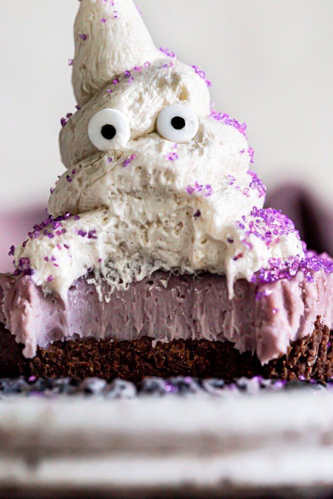 vegan cheese cake with a chocolate crust and purple vegan cashew cheesecake layer topped with a buttercream frosting ghost with candy eyes and purple sprinkles.