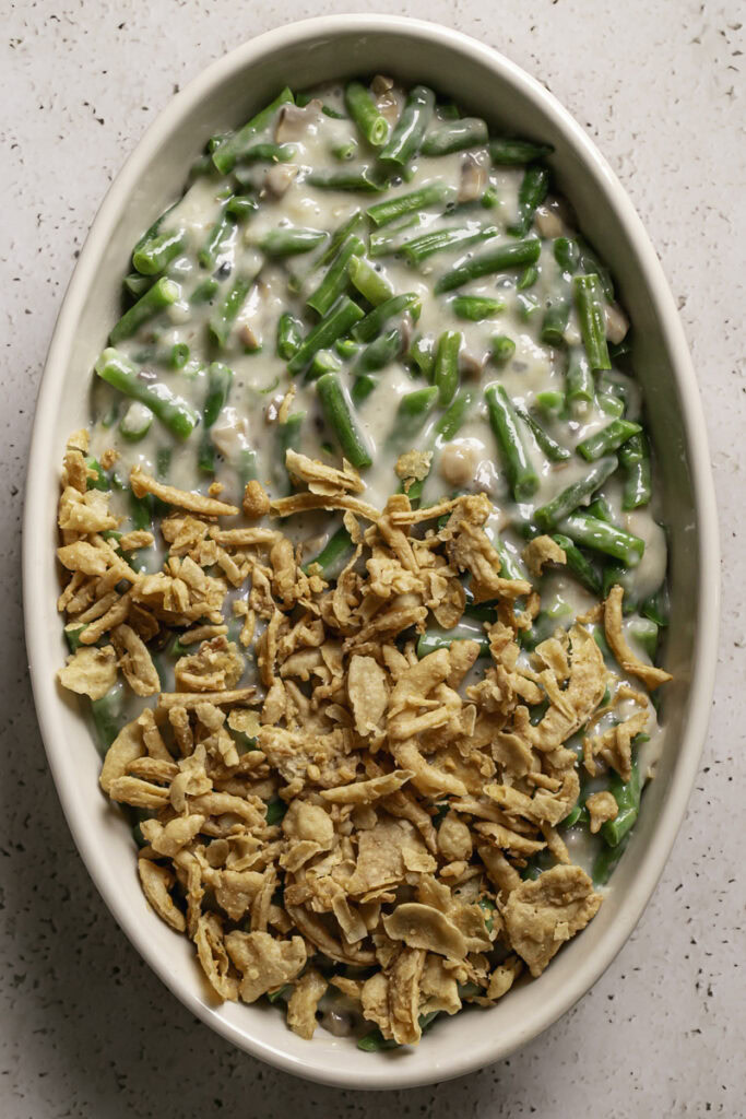 an oval casserole dish filled with uncooked green bean casserole and half topped with crispy fried French onions