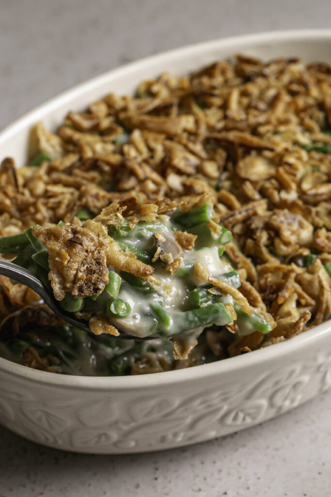 a close up of a spoon scooping some green bean casserole topped with crispy fried French onions out of a casserole dish