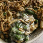 a close up of a spoon scooping some green bean casserole topped with crispy fried French onions out of a casserole dish