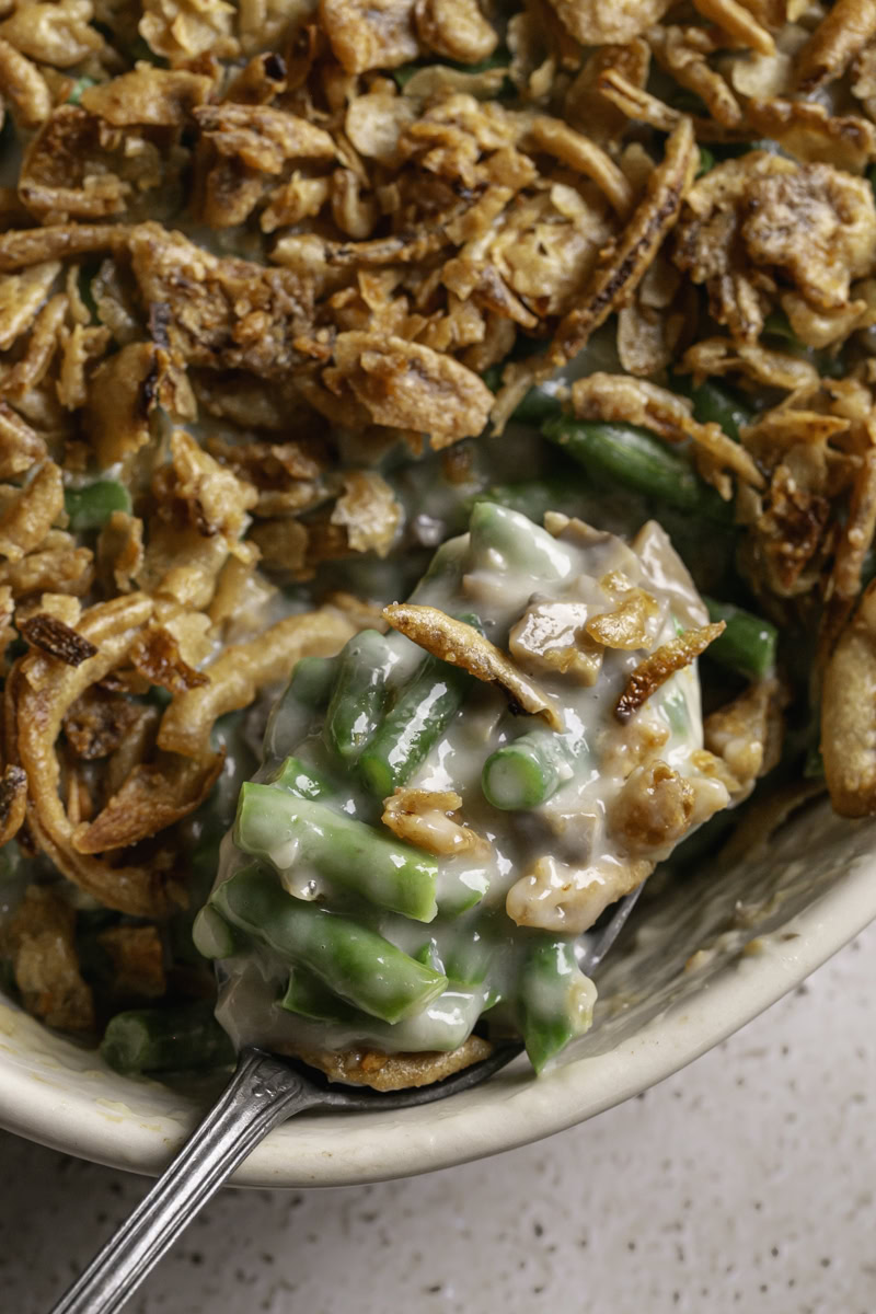 a close up of a spoon scooping some green bean casserole topped with crispy fried French onions out of a casserole dish