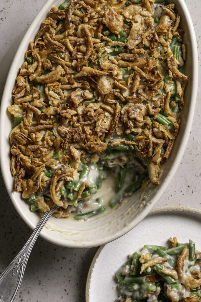 an oval casserole dish with green bean casserole, some scooped out, a serving spoon in the casserole dish and the scooped out casserole on a plate
