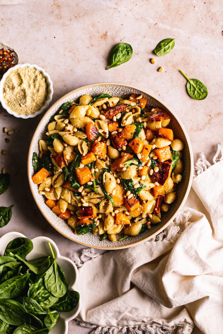 a bowl of pasta tossed with roasted butternut squash and spinach and garnished with red pepper flakes, vegan parmesan cheese, and toasted pine nuts. A bowl of fresh spinach on the side and small bowls of vegan parmesan cheese and red pepper flakes and a cream colored napkin