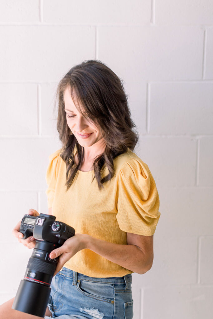 Gina Fontana sitting on a stool wearing a yellow top looking down at her canon camera to preview images she took
