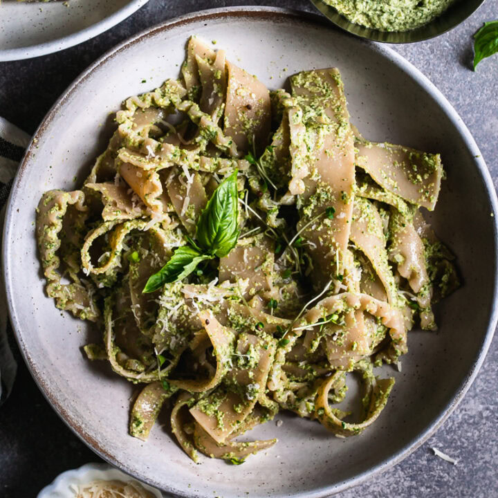a close up of a bowl of olive pesto pasta garnished with a basil leaf a bowl of olive pesto and vegan parmesan cheese beside the bowl of pasta