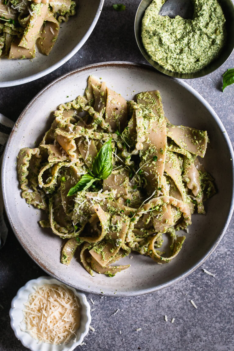 a close up of a bowl of olive pesto pasta garnished with a basil leaf a bowl of olive pesto and vegan parmesan cheese beside the bowl of pasta