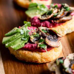 close up of hash browns topped with beet hummus, sliced avocado, balsamic roasted mushrooms, vegan parmesan cheese and micro greens