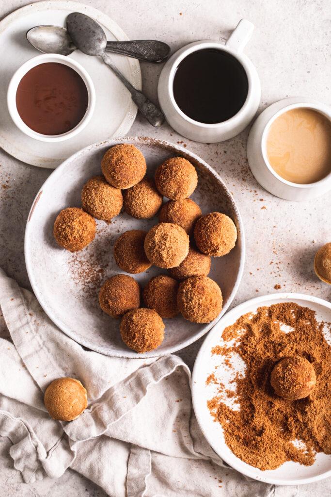 a bowl of grain-free, gluten-free, vegan, naturally sweetened churro donut holes with a bowl of cinnamon coconut sugar beside it with a donut hole in it, a bowl of melted chocolate for dipping and two mugs of coffee