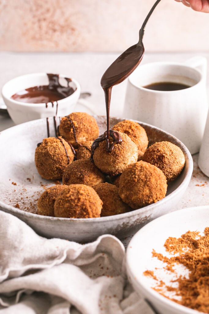 a side view of gluten-free, grain-free, vegan, naturally sweetened churro donut holes with a mug of coffee and a bowl full of melted chocolate in the background and a drizzle of chocolate from a spoon on one of the donut holes
