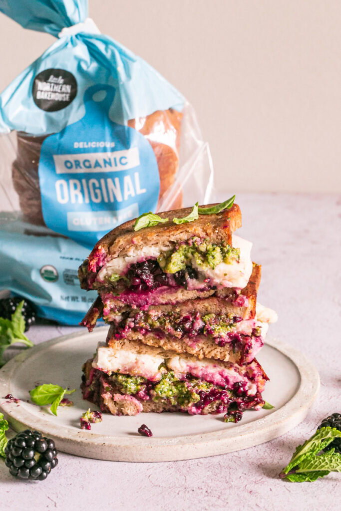 vertical image of vegan blackberry pesto grilled cheese sandwich stacked on top of one another and a loaf of little northern bakehouse bread in the background