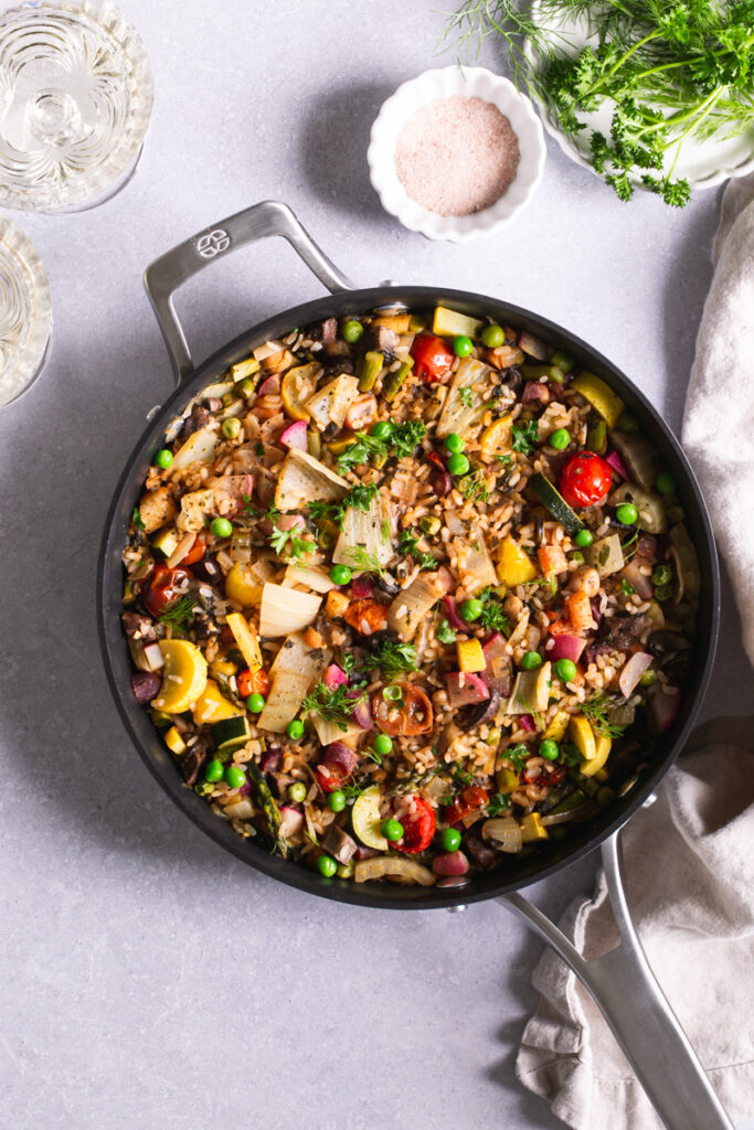 calphalon saute pan with prepared vegetable paella a bowl of salt, plate of herbs, and two glasses of wine next to it