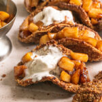 3/4 image of apple pie tacos with partially melted ice cream on top and a bowl of apple pie filling in the background