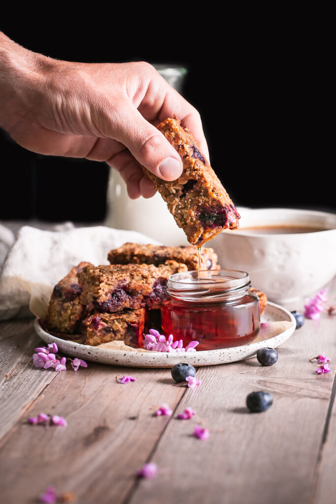 Blueberry Oatmeal French Toast Sticks on a plate one in hand being dipped in a jar of maple syrup with a jar of maple syrup beside it and a cup of coffee and jug of almond milk in the background