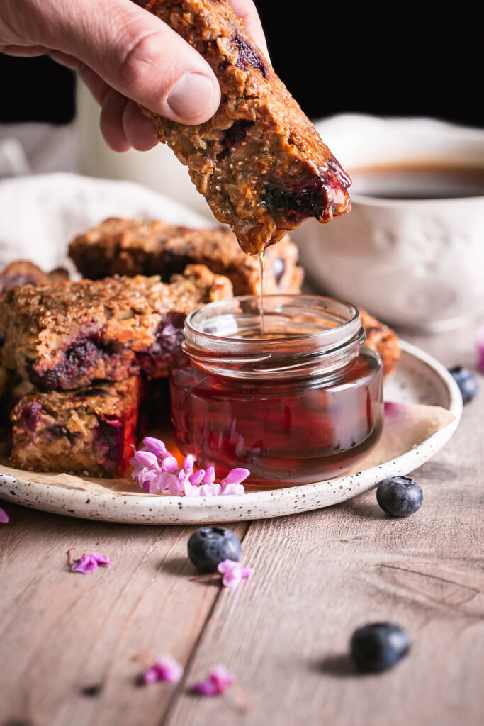 close up of Blueberry Oatmeal French Toast Sticks on a plate one in hand being dipped in a jar of maple syrup with a jar of maple syrup beside it and a cup of coffee and jug of almond milk in the background