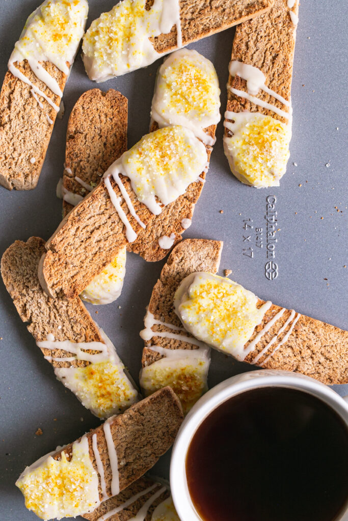 lemon vanilla biscotti piled on top of each other on a Calphalon cookie sheet with a cup of coffee