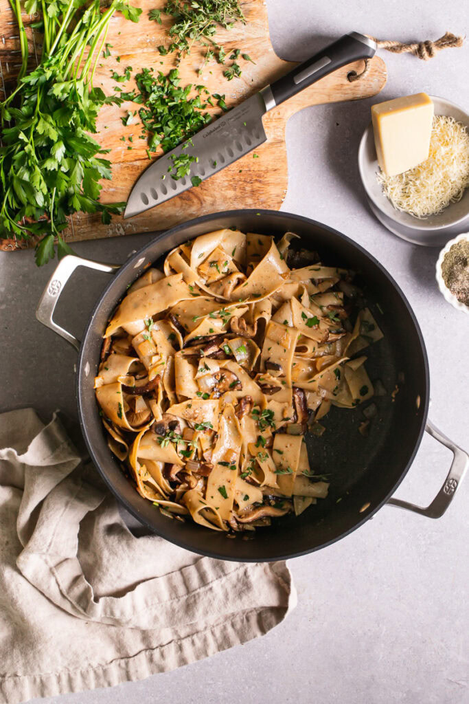a pot of Gluten-Free Pappardelle with Wild Mushroom Ragù with a cutting board above it with fresh parsley and thyme and a knife, a plate of vegan parmesan cheese and a bowl of salt and pepper.
