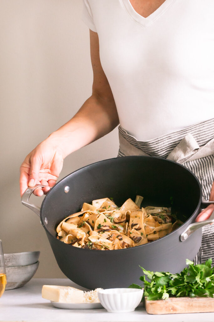 a woman holding a pot of Gluten-Free Pappardelle with Wild Mushroom Ragù with a cutting board with parsley on it, vegan parmesan cheese in front