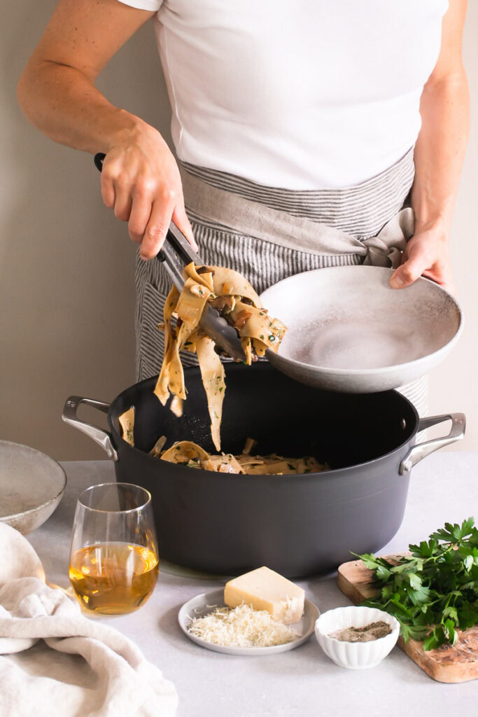 a woman with a pot of Gluten-Free Pappardelle with Wild Mushroom Ragù serving some into a bowl with tongs.