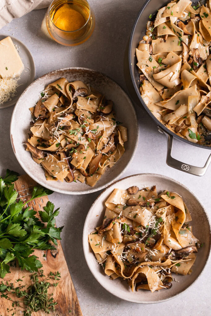 two bowls of gluten-free pappardelle with wild mushroom Ragù next to the serving pot of pasta, a glass of wine, and a cutting board with fresh parsley beside it