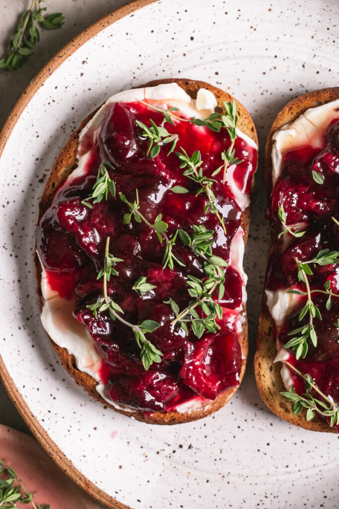 a close up shot of two slices of toast with dairy-free cream cheese, roasted cherries and fresh thyme on a plate