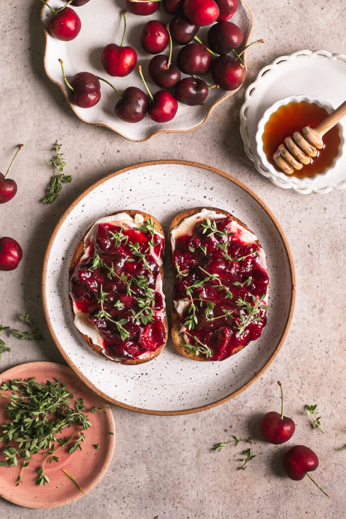 two slices of toast with dairy-free cream cheese, roasted cherries and fresh thyme on top and a plate of fresh thyme, a plate of fresh cherries, and a bowl of agave syrup around it
