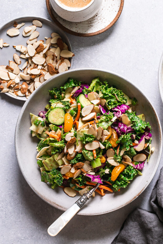 a bowl of Thai chopped salad with a fork in the bowl, a plate of sliced almonds and dressing next to the bowl