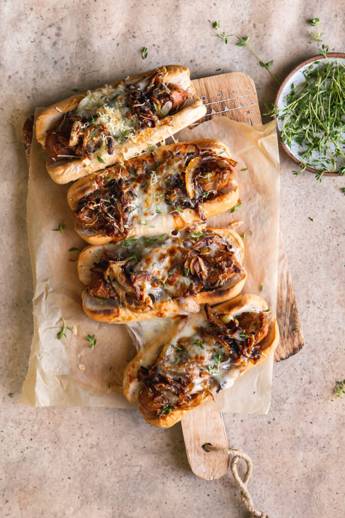 four vegan French onion brats layed out on a parchment line wooden cutting board with a bowl of fresh thyme next to it