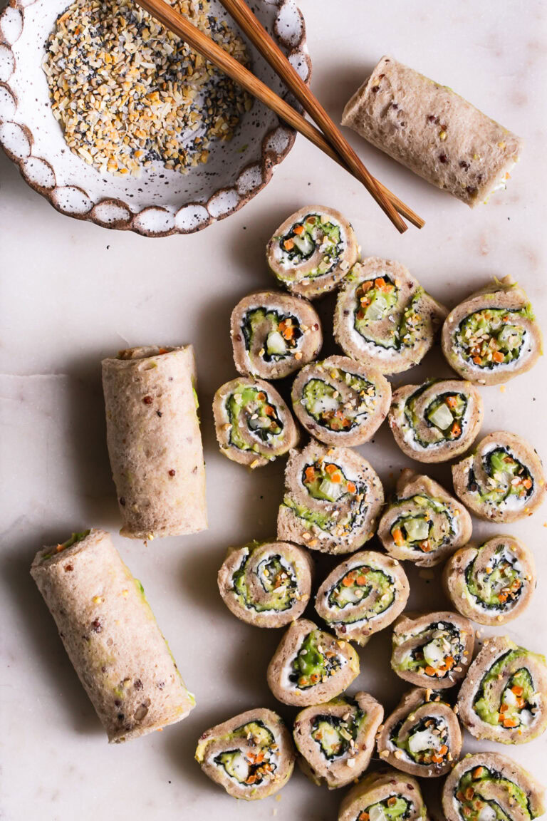 a marble cutting board with 3 rolled up unsliced veggie sushi sandwiches and sliced rollup sandwiches with a bowl of everything bagel spice and wooden chopsticks