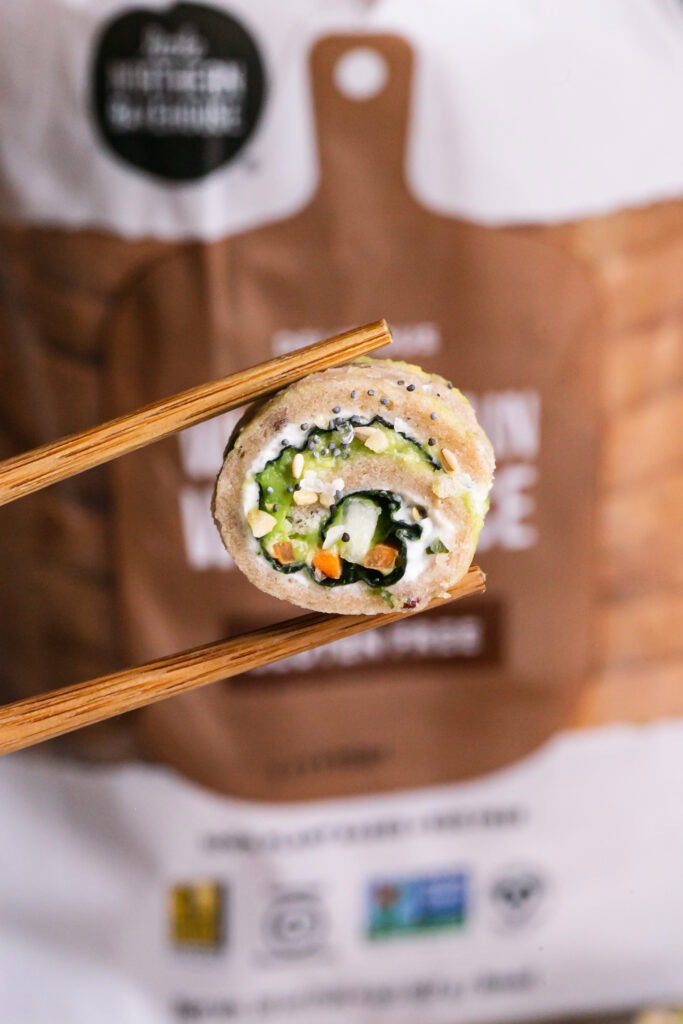 close up vertical image of a slice of veggie sandwich sushi held with chopsticks and a loaf of Little Northern Bakehouse whole grain bread in the background