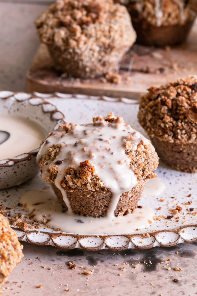 3/4 shot of a frosted cinnamon streusel muffin with a bowl of icing beside it on a plate