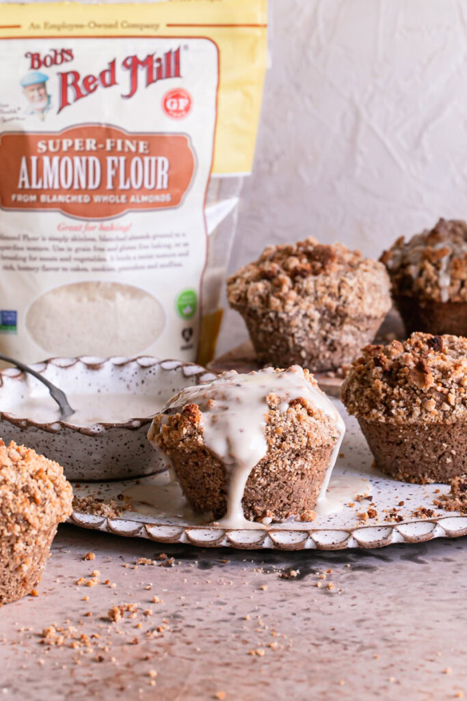 one iced cinnamon streusel muffin with other unfrosted muffins around it, a bowl of icing beside it and a bag of Bob's Red Mill blanced almond flour in the background